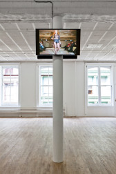 Two screens hung on either side of the top of a support beam in a gallery. The room is otherwise empty. One screen depicts a woman walking down a fashion runway, wearing a black jacket and shorts with a sheer top.