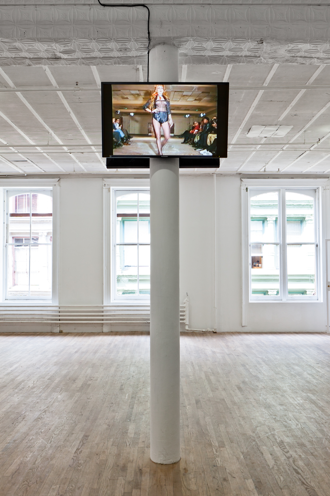Two screens hung on either side of the top of a support beam in a gallery. The room is otherwise empty. One screen depicts a woman walking down a fashion runway, wearing a black jacket and shorts with a sheer top.