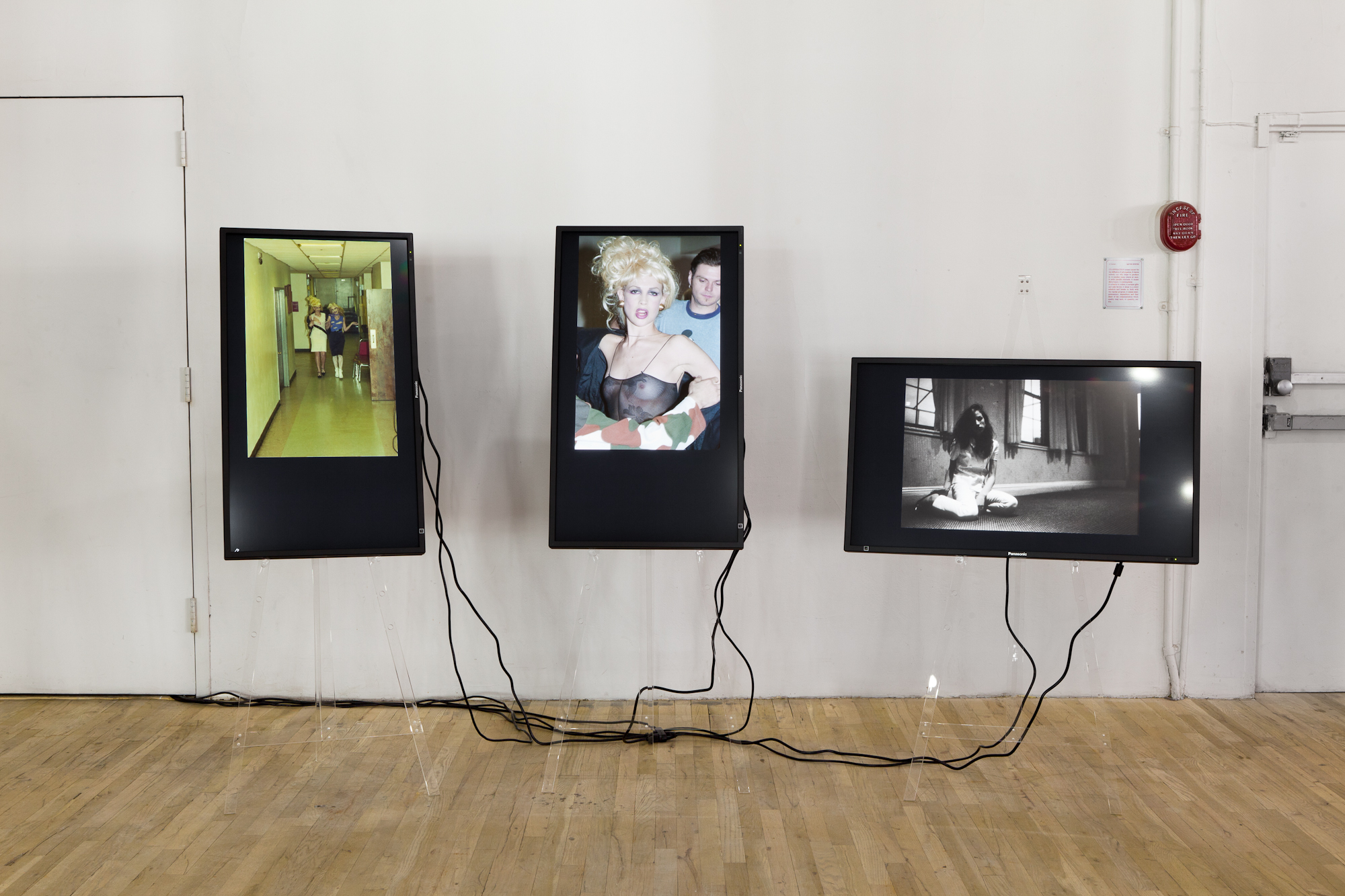 Three television screens installed on a gallery wall. Two screens are positioned in a portrait orientation and the third in a landscape orientation. Their cords loosely fall to the ground below and run beneath a door. Each screen displays images of one or several figures mid-speech or mid-movement.
