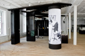 A large, shiny black structure consisting of three columns supporting a large, irregularly shaped flat plane installed in a gallery space. A black-and-white image wraps around the rightmost, cylindrical column of the structure. The image depicts a person dressing a mannequin in an office.
