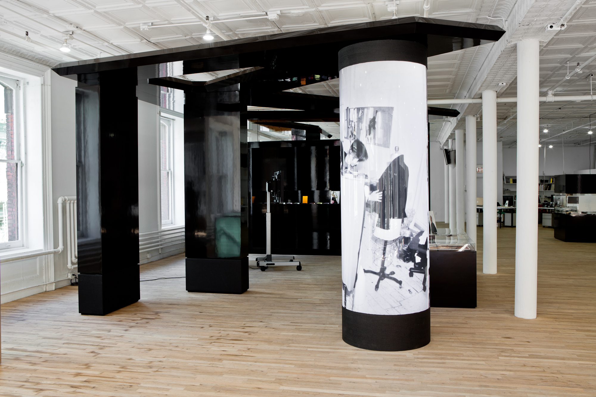 A large, shiny black structure consisting of three columns supporting a large, irregularly shaped flat plane installed in a gallery space. A black-and-white image wraps around the rightmost, cylindrical column of the structure. The image depicts a person dressing a mannequin in an office.
