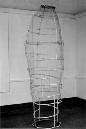 A black and white photograph of a sculpture, which sits on the ground near the corner of a room, its wiry frame covered by a transparent mesh material.