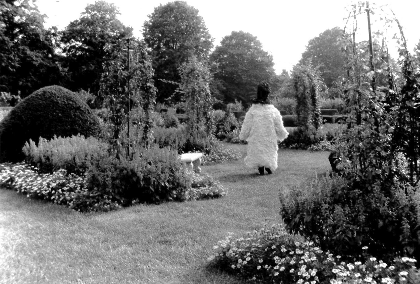 A photograph of a figure in a long furry coat who walks along a path in a lush garden, their back facing the camera.