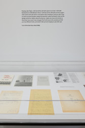 A close up image of a glass display case holding, images, books, and handwritten text. On the wall above is a paragraph of text that reads, 