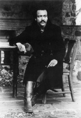 A person sits on a wooden chair and looks off into the distance. His right elbow rests on a table partially visible in the photograph.