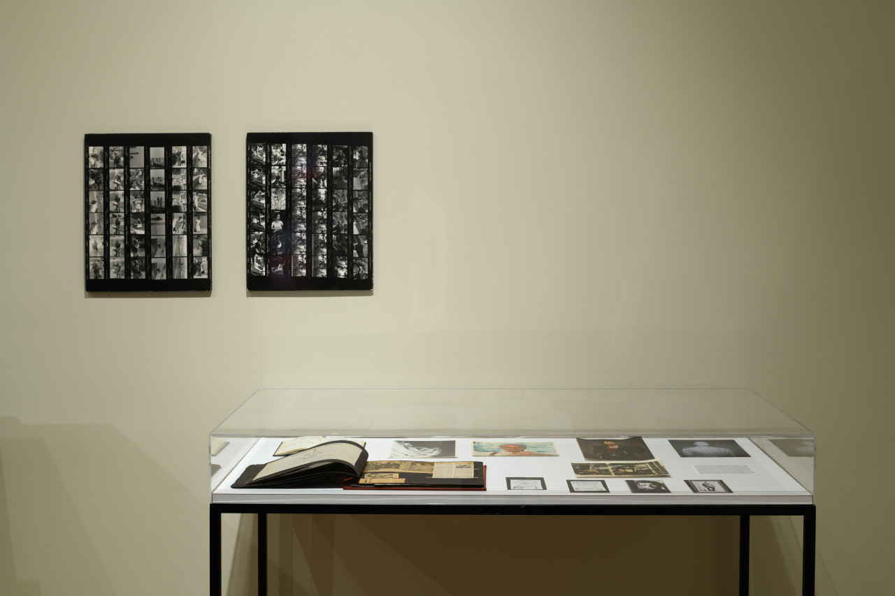 Along the wall is a glass display case holding ephemera, hanging above are two black and white contact sheets.