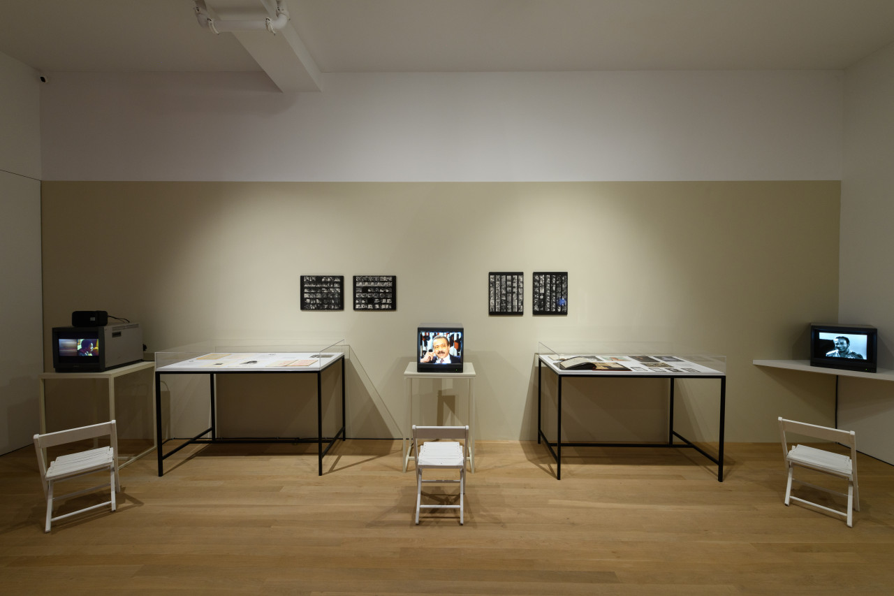 A wide angle image of a beige wall lined with multiple tv monitors and glass display cases filled with ephemera, hanging on the wall are four black and white contact sheets.