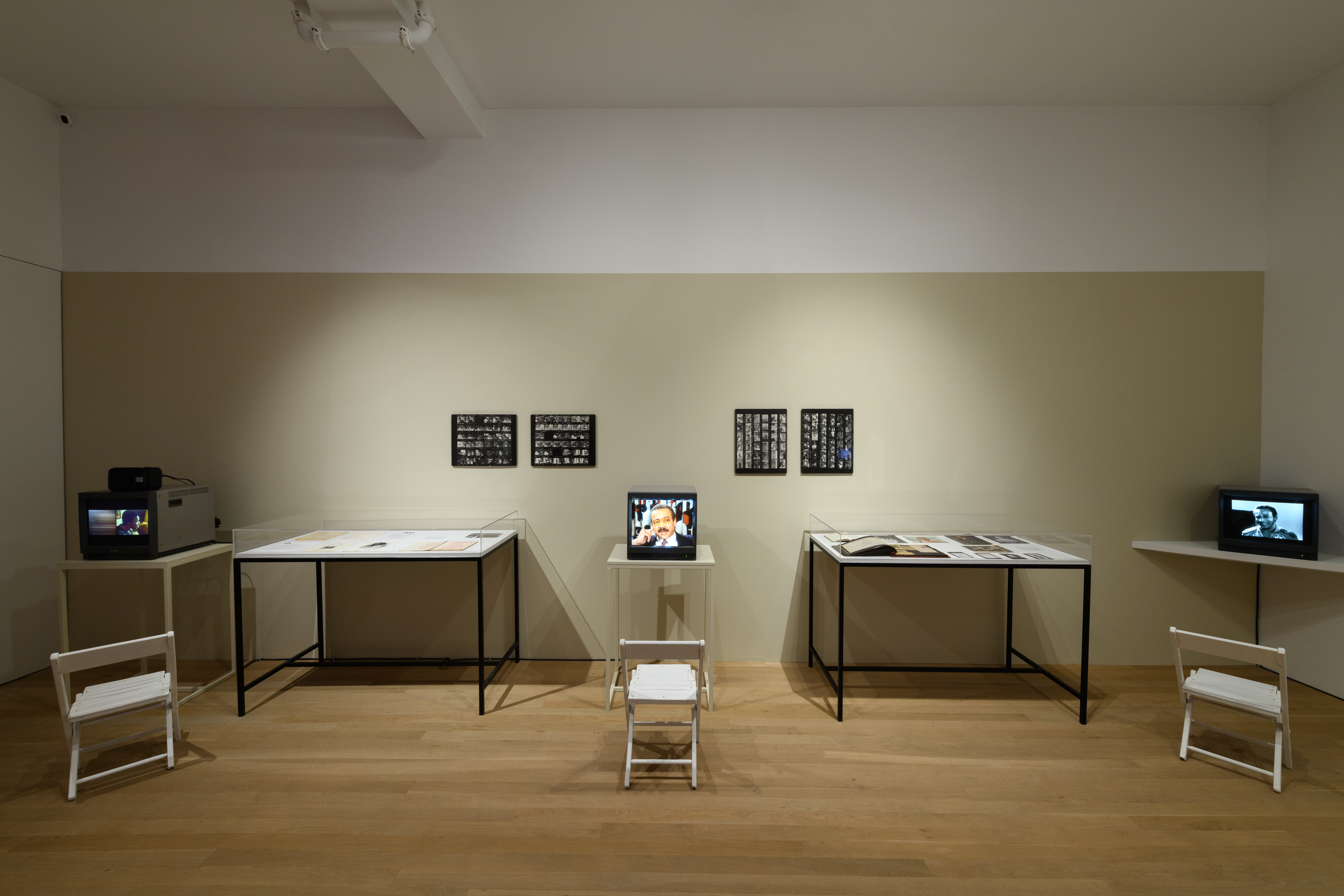 A wide angle image of a beige wall lined with multiple tv monitors and glass display cases filled with ephemera, hanging on the wall are four black and white contact sheets.