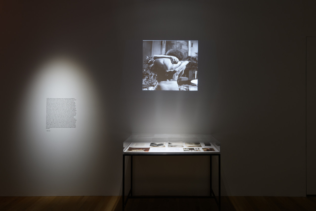 An image of a dark room with two spotlights, one pointed at wall text, and the other at a wall projection and glass display case.