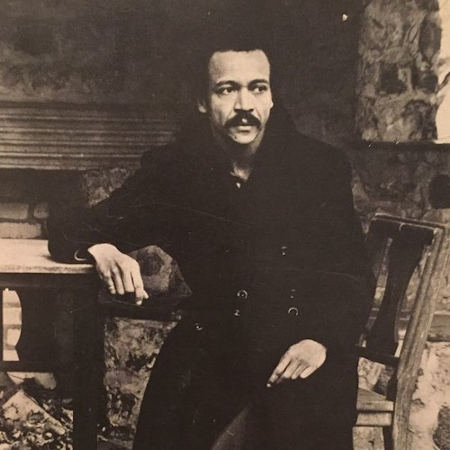 A person sits on a wooden chair and looks off into the distance. His right elbow rests on a table partially visible in this sepia-toned photograph.