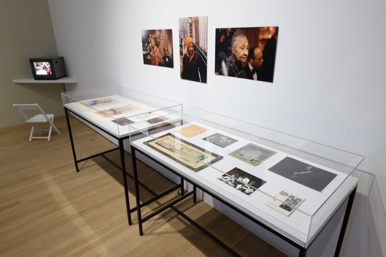 Along the wall are two glass display cases holding ephemera, in the center of them three images hang on the wall, on the left sits a tv monitor on a shelf.