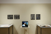 Two glass display cases holding ephemera sit on either side of a tv monitor, on the wall are four black and white contact sheets.