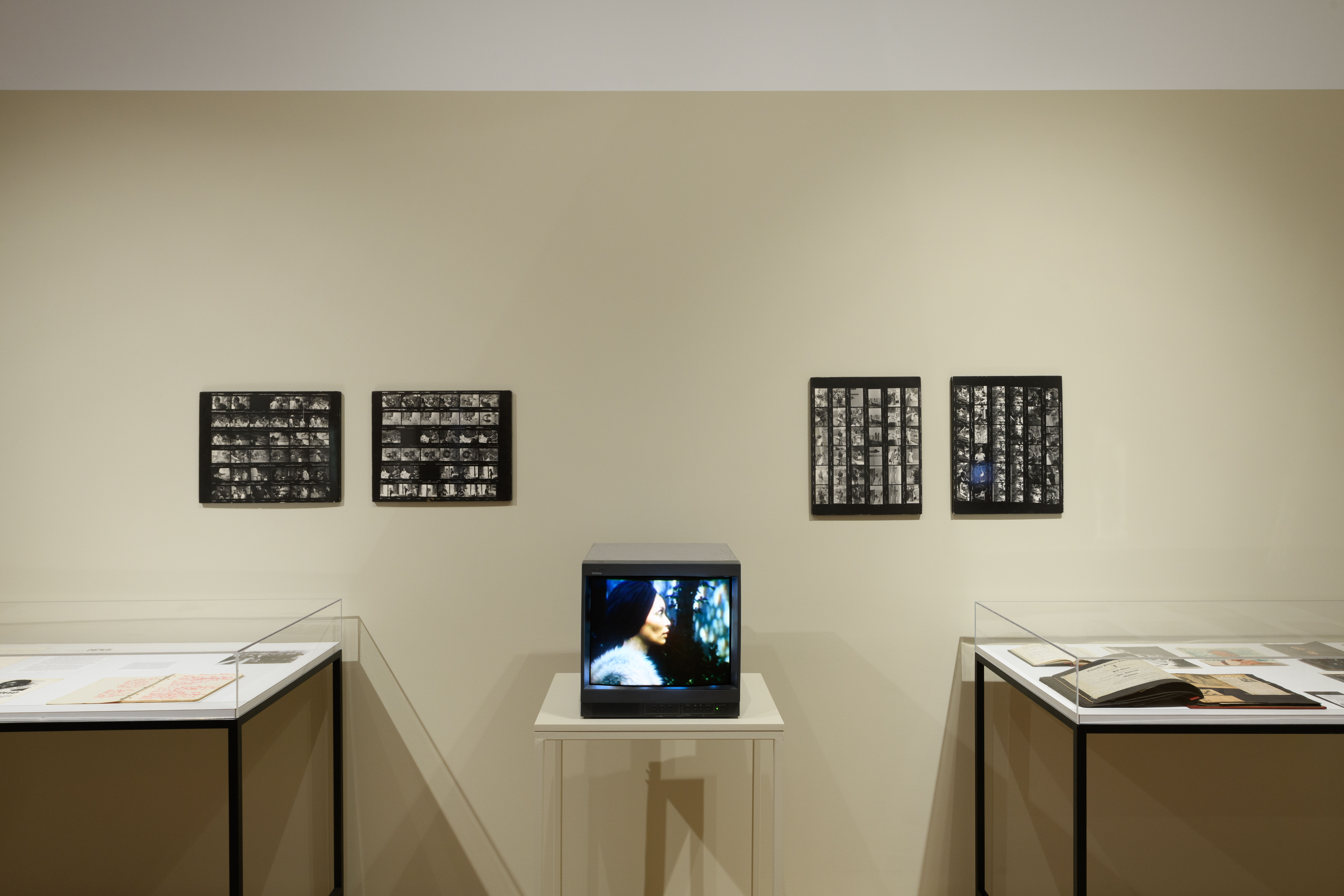 Two glass display cases holding ephemera sit on either side of a tv monitor, on the wall are four black and white contact sheets.