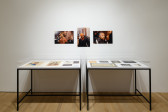Along the wall are two glass display cases filled with ephemera, in the center of them, three images hang on the wall.