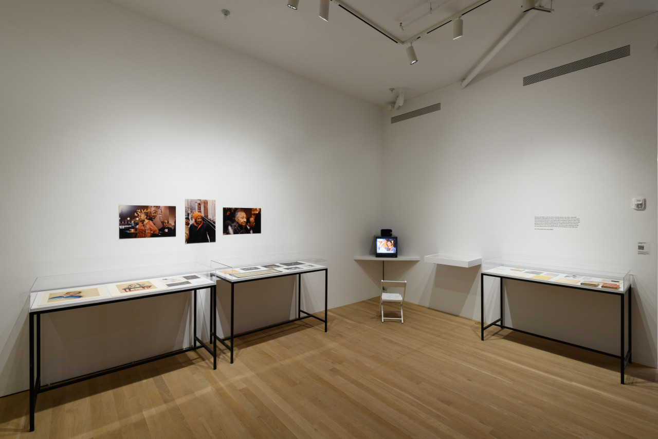 Lining the wall are multiple glass display cased filled with ephemera, text and images hang on the wall above them, in the corner sits a tv monitor on a shelf.