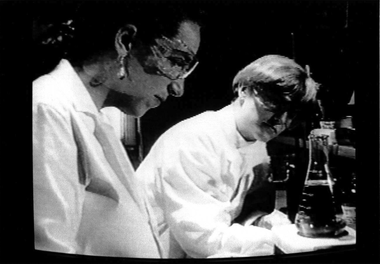 Two people in lab coats and protective goggles are shown in black and white, studying a beaker on the table in front of them.