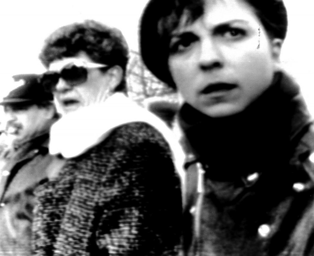 A black and white image of three people wearing winter coats and scarves, observing something to the left of the camera.