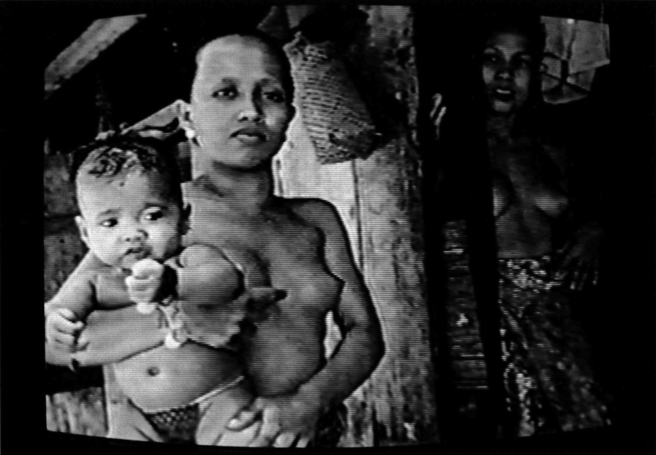 A black and white image of a shirtless woman holding a baby, while another woman looks on from a doorway to the right.