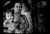 A black and white image of a shirtless woman holding a baby, while another woman looks on from a doorway to the right.