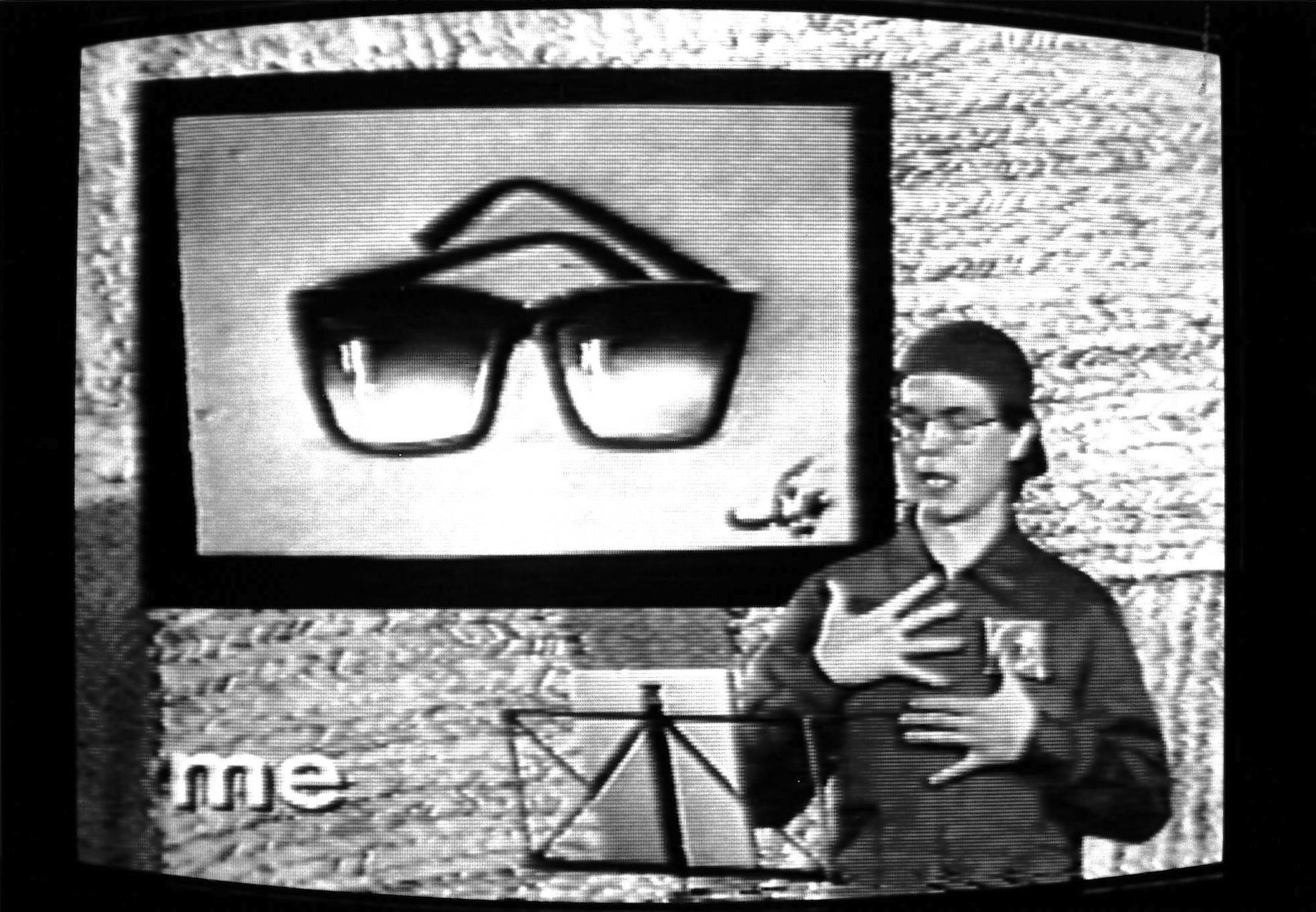 A black and white image of person in a coverall jumpsuit and backwards baseball cap reading aloud from a music stand.  Behind them, a pair of sunglasses is shown on a white background.