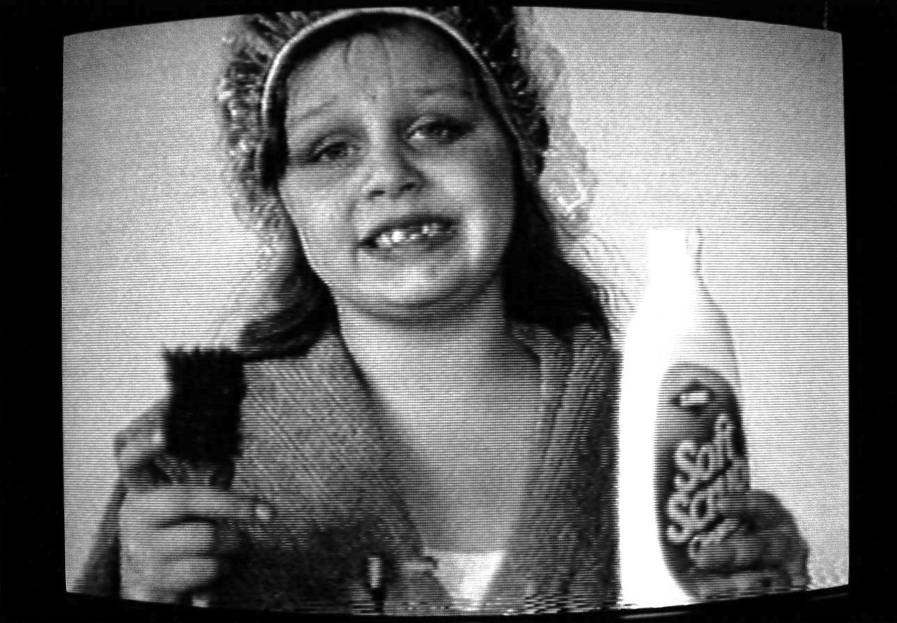 A black and white close-up image of an adolescent girl, smiling at the camera.  She is holding up a scrub brush in one hand and a bottle of Soft Scrub in the other and is wearing a bathrobe and a shower cap.