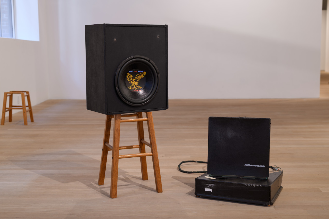 A large speaker rests on a small wooden stool. To the right sit two abstracted black box-like objects.