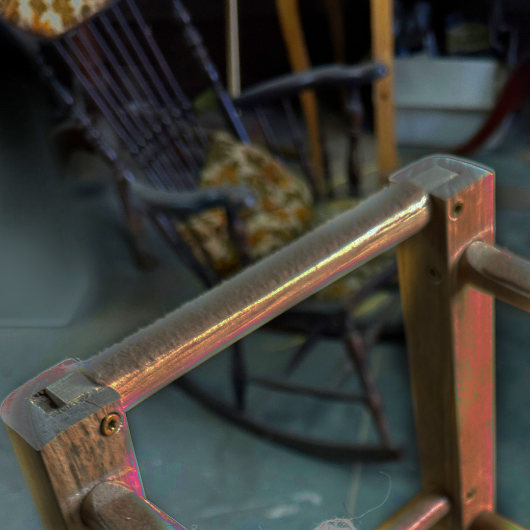 An upside-down wooden stool is cast in a glowing, neon light. In the background, a rocking chair with decorative pillows appears out of focus.