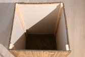 An overhead shot depicts the interior of a wooden box-like structure. The floor of the object is lined with dark carpet, and the walls of the structure bear a cream geometric pattern. 