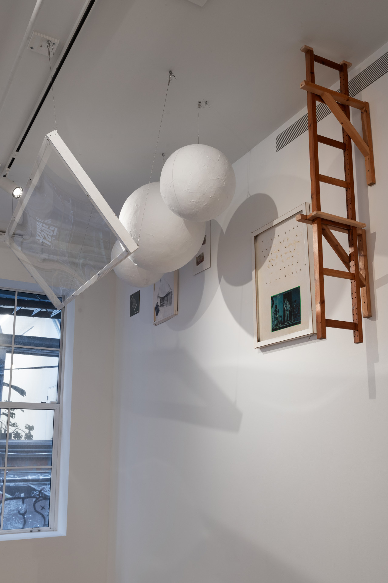 A transparent sheet suspends from the ceiling, framing three large white balls and a selection of drawings. Protruding from the wall is a wooden ladder.