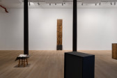 Two iron columns run across the gallery floor. Central in the room, on the back wall, five rectangular boards stand stacked on top of each other. The bottom plank is darker in color. In front of the columns, a stool adorned with a box sits to the left. Two abstracted boxes sit to the right, one black, the other made of a wood material similar to the central stack. 