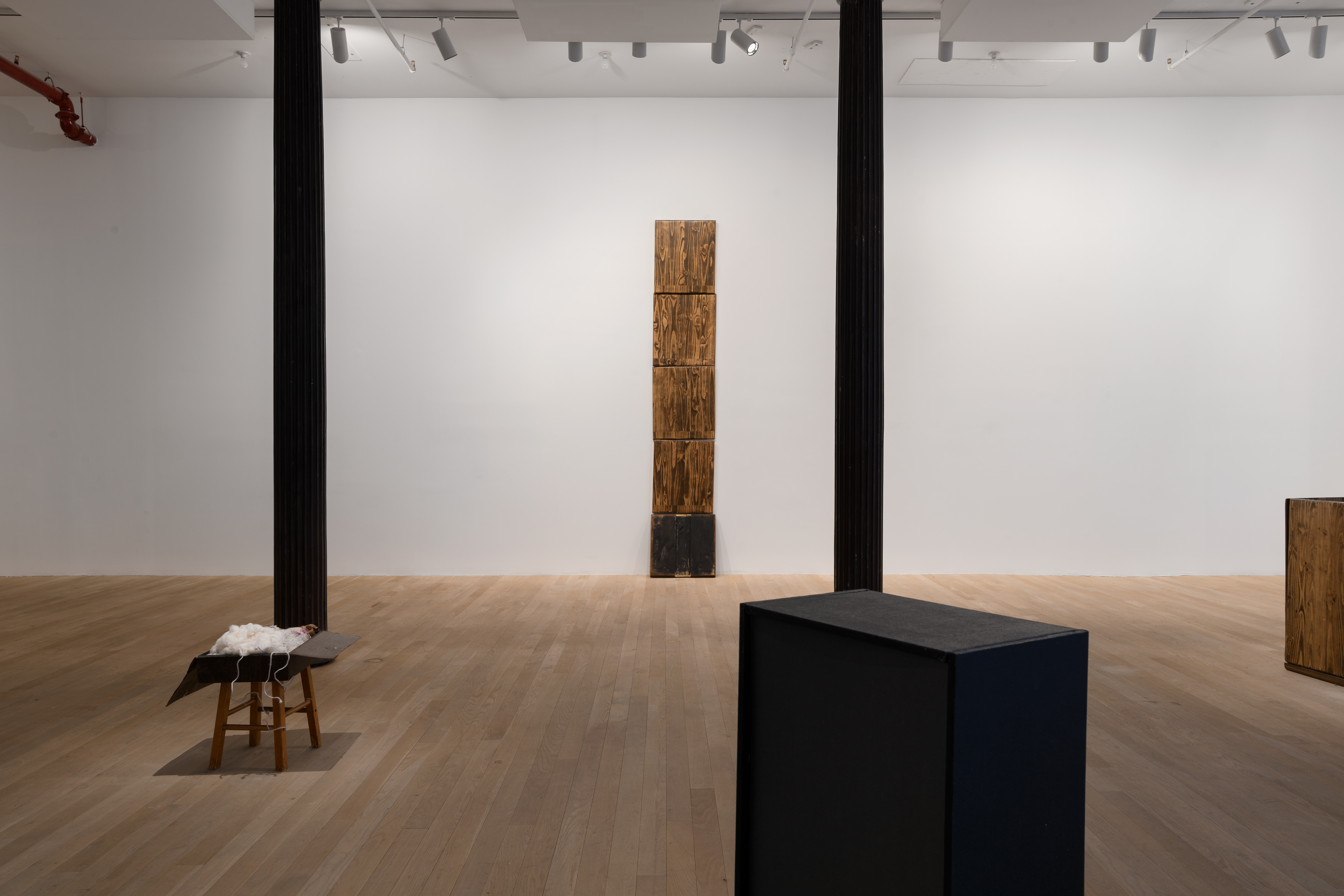 Two iron columns run across the gallery floor. Central in the room, on the back wall, five rectangular boards stand stacked on top of each other. The bottom plank is darker in color. In front of the columns, a stool adorned with a box sits to the left. Two abstracted boxes sit to the right, one black, the other made of a wood material similar to the central stack. 