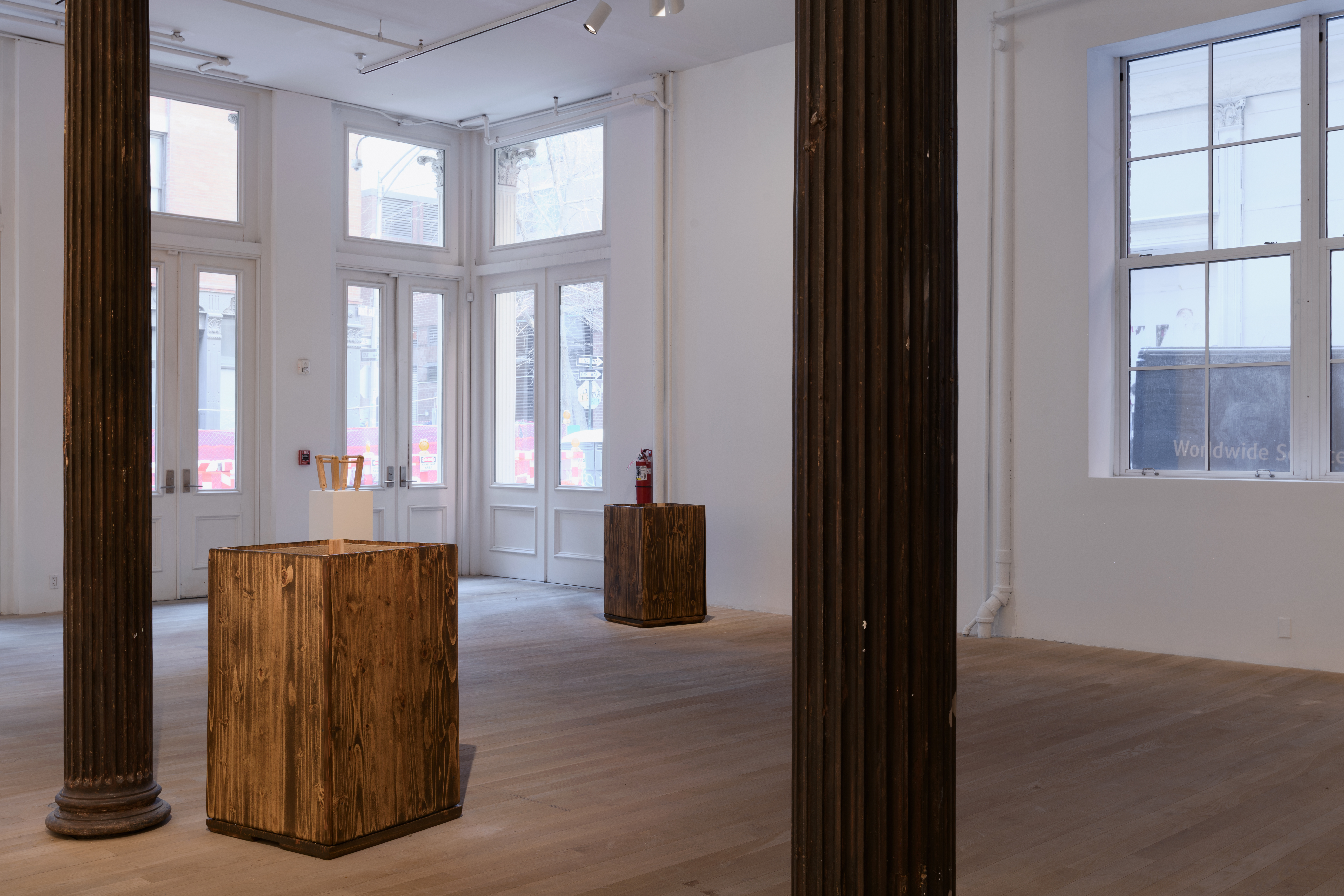 Placed at an angle between two iron columns are two box-like structures made of wooden boards. Behind the closer box, a pedestal presents a small wooden stool. 