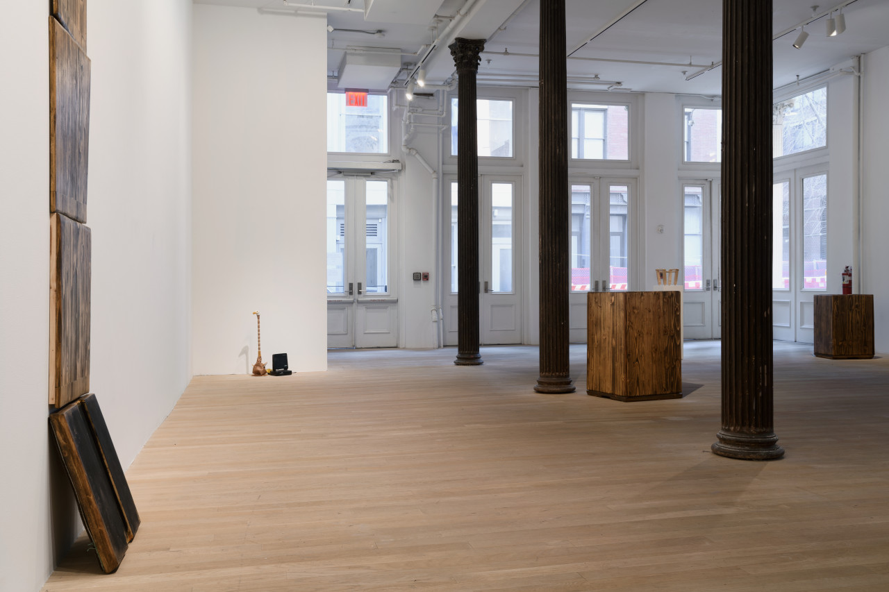 Stacked wooden planks stand on the left wall. One plank rests at an angle as if supporting the others above. Three other sculptures are seen dispersed around the gallery.