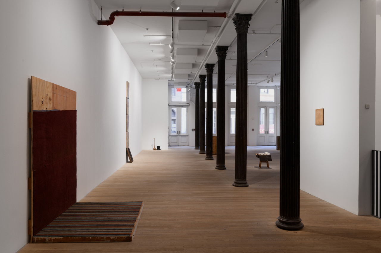 Iron columns run down the center of the open gallery. To the left sits a pair of wooden boards adorned with maroon and striped carpet. On the wall opposite hangs a smaller wooden square. A stool-like structure stands to the right of the iron columns.