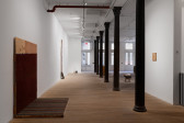 Iron columns run down the center of the open gallery. To the left sits a pair of wooden boards adorned with maroon and striped carpet. On the wall opposite hangs a smaller wooden square. A stool-like structure stands to the right of the iron columns. 