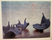 Three dark-colored objects are arranged on a wooden surface. The solid, organic forms curl and fold up towards their portruding tops.