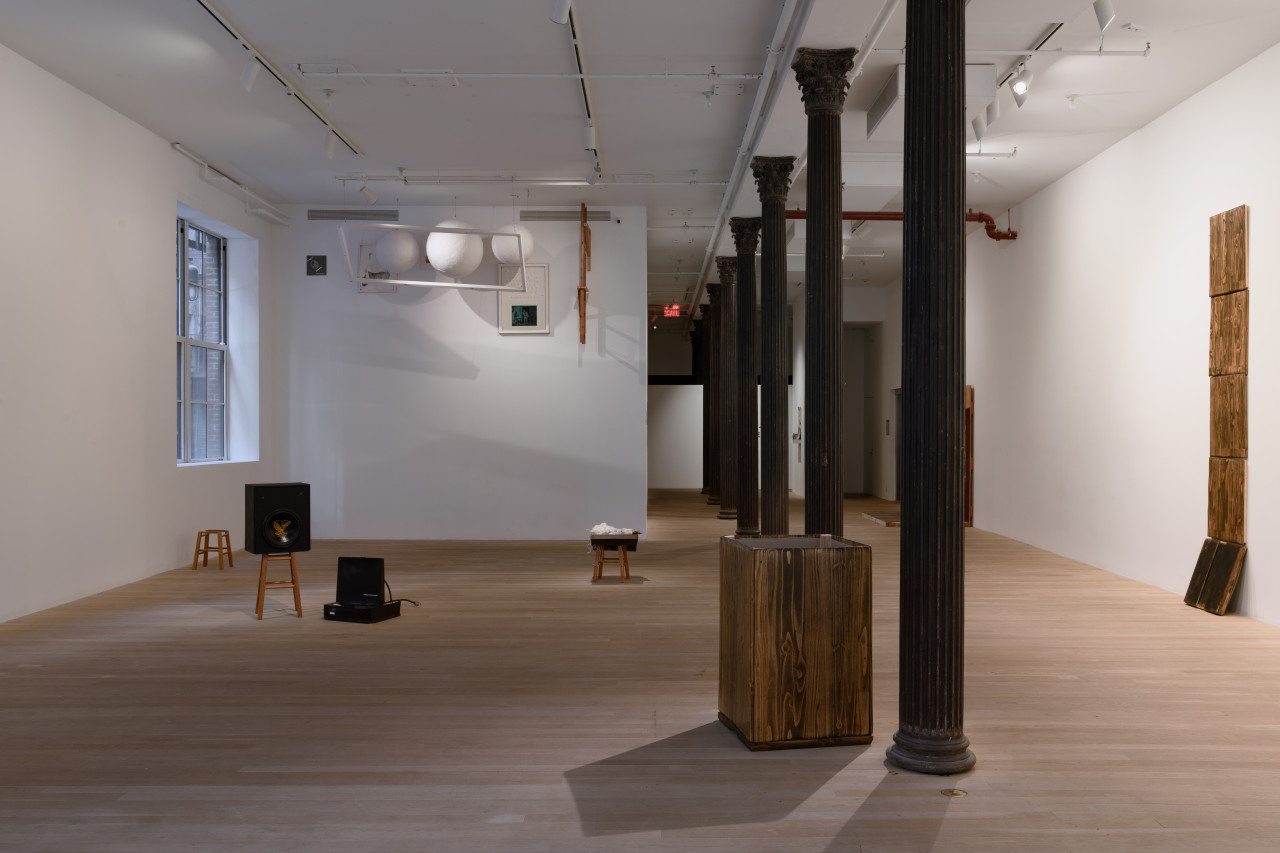 To the left, various stools and black boxes are dispersed around the floor of the gallery. Suspended above hangs a transparent sheet, framing three large white balls and a selection of drawings. Wooden rectangular panels stand stacked on the right wall. Towards the center of the space is a box-like structure made of similar wooden paneling.