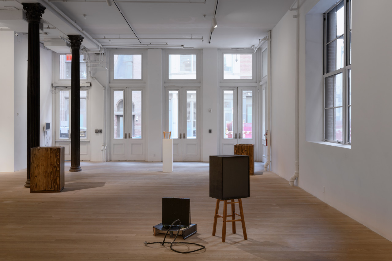 Various box-like structures fill the gallery space. Central in the space, a white pedestal presents a small wooden stool.
