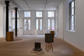 Various box-like structures fill the gallery space. Central in the space, a white pedestal presents a small wooden stool. 