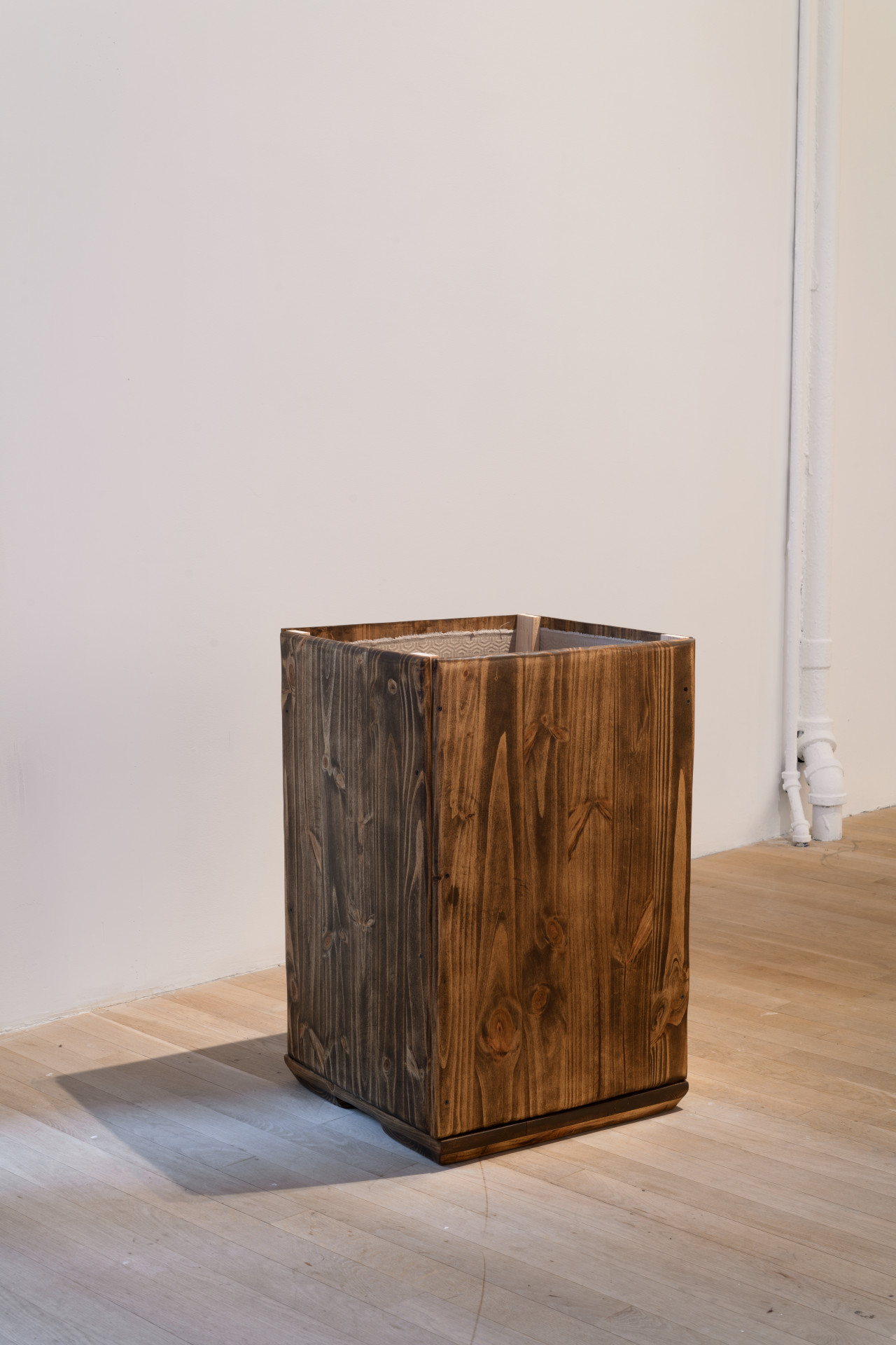 A box-like structure made of wooden boards casts a shadow. The top of the structure is open, revealing an interior lining.