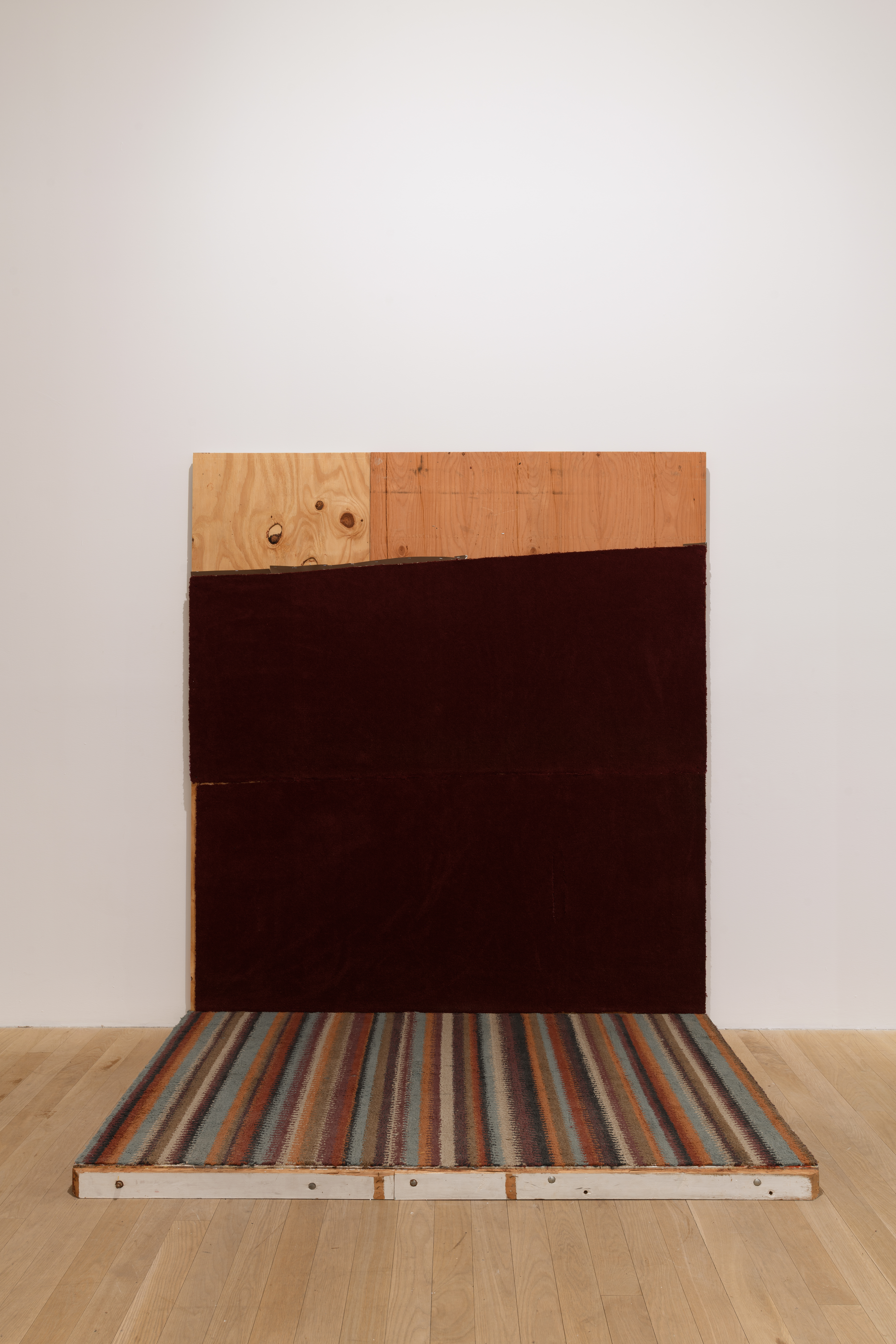 Two boards, one horizontal on the ground and the other vertical on the wall, meet at a right angle. The bottom board sits covered with stripped carpet, the top nearly covered with marroon carpet. 