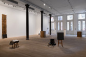 An open gallery presents various works dispersed throughout the space. To the far left, five rectangular wooden boards stand stacked on top of each other. The bottom board rests at an angle as if to support the others above. 