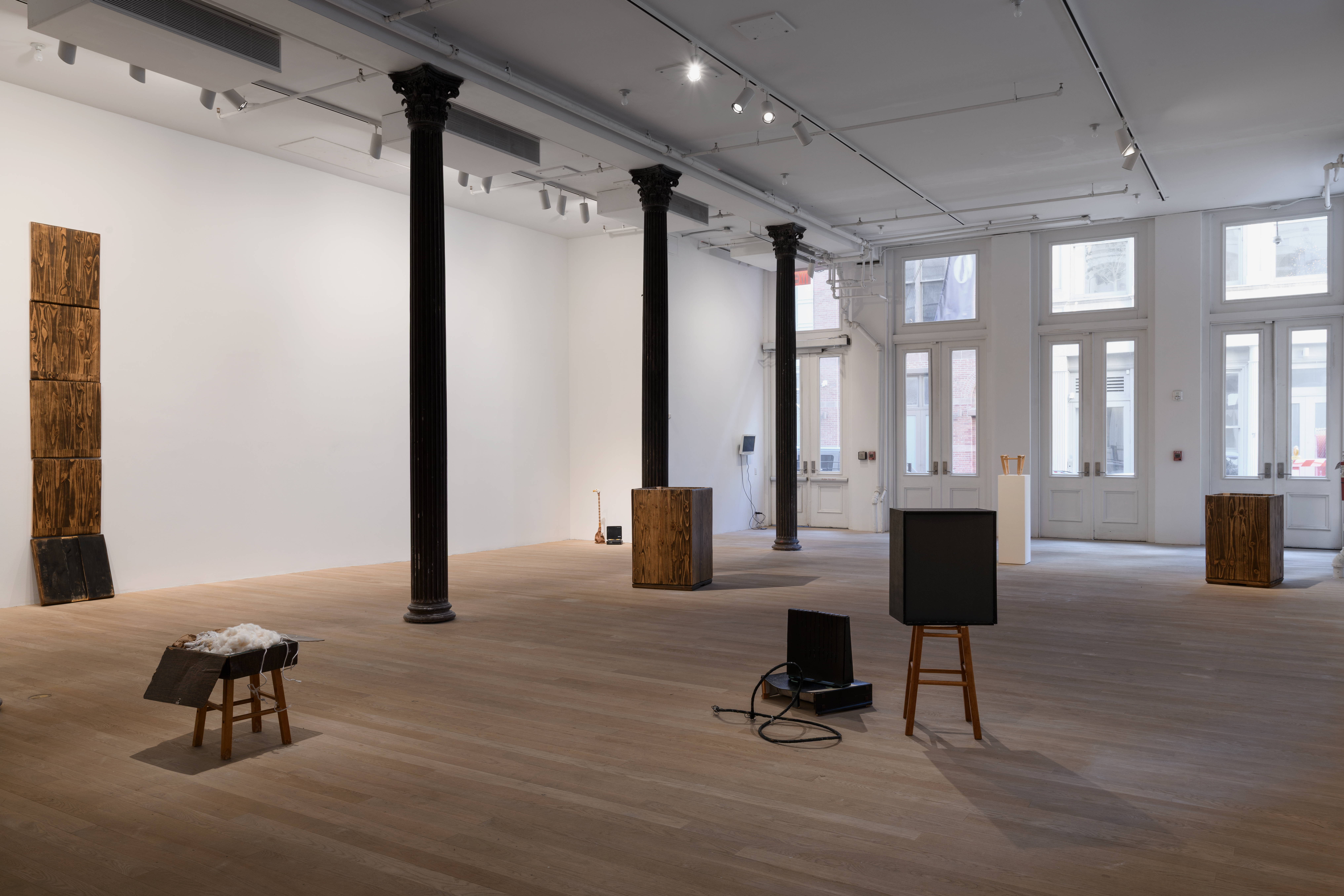 An open gallery presents various works dispersed throughout the space. To the far left, five rectangular wooden boards stand stacked on top of each other. The bottom board rests at an angle as if to support the others above. 