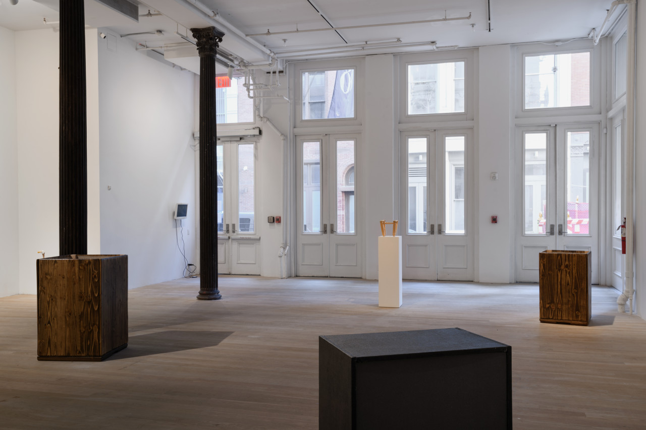 Placed at an angle from each other stand two box-like structures made of wooden boards, a black speaker, and a white pedestal bearing a small wooden stool.