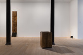 Three works are seen in the gallery space. To the far left, five rectangular wooden boards stand stacked on top of each other. Central is a tall box made of similar wood boards. To the right sits an eagle-headed sculpture and an object resembling a modem. 
