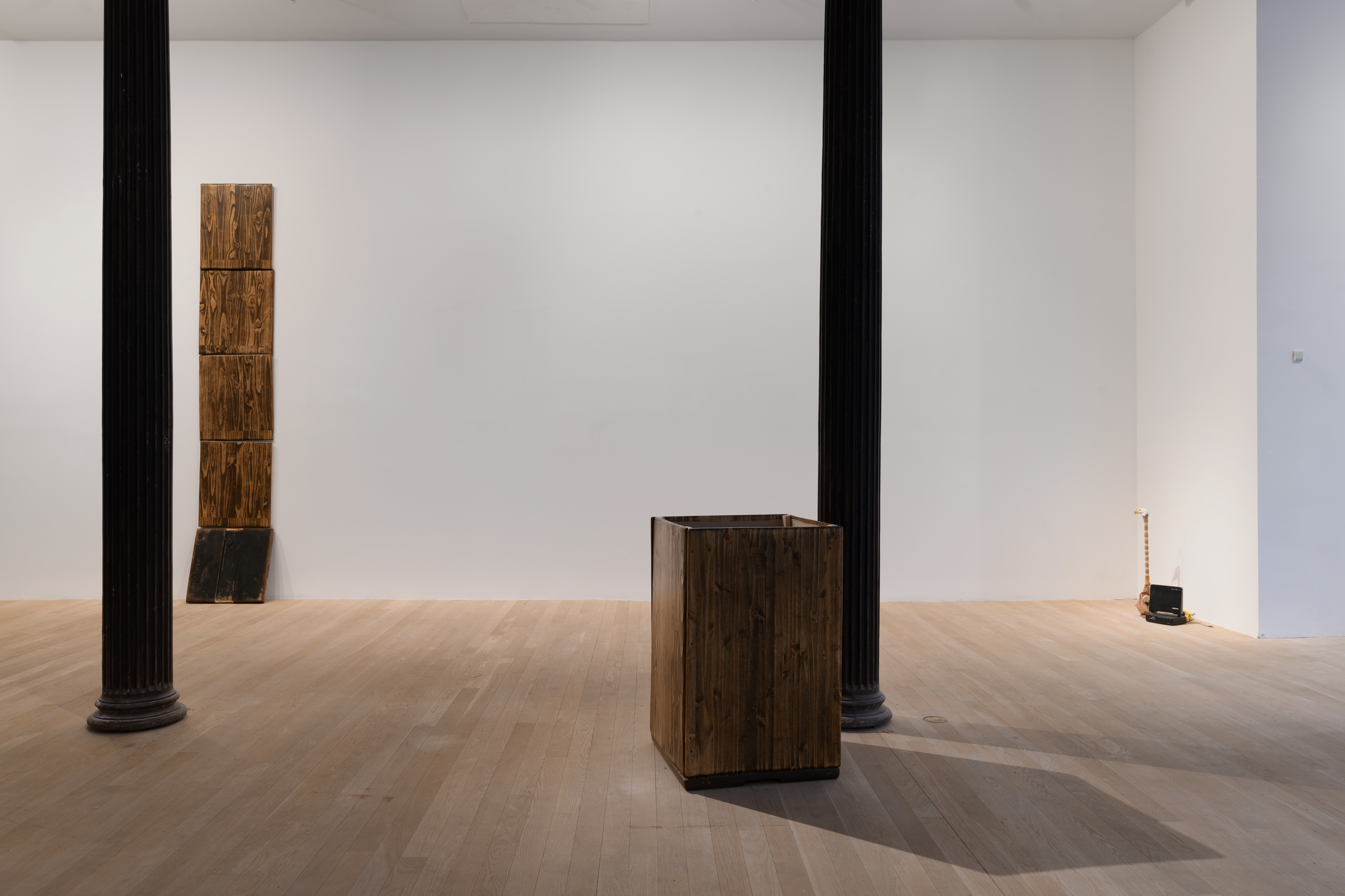 Three works are seen in the gallery space. To the far left, five rectangular wooden boards stand stacked on top of each other. Central is a tall box made of similar wood boards. To the right sits an eagle-headed sculpture and an object resembling a modem. 