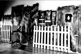 Black and white photograph of white picket fence opening with dark painted background behind it.