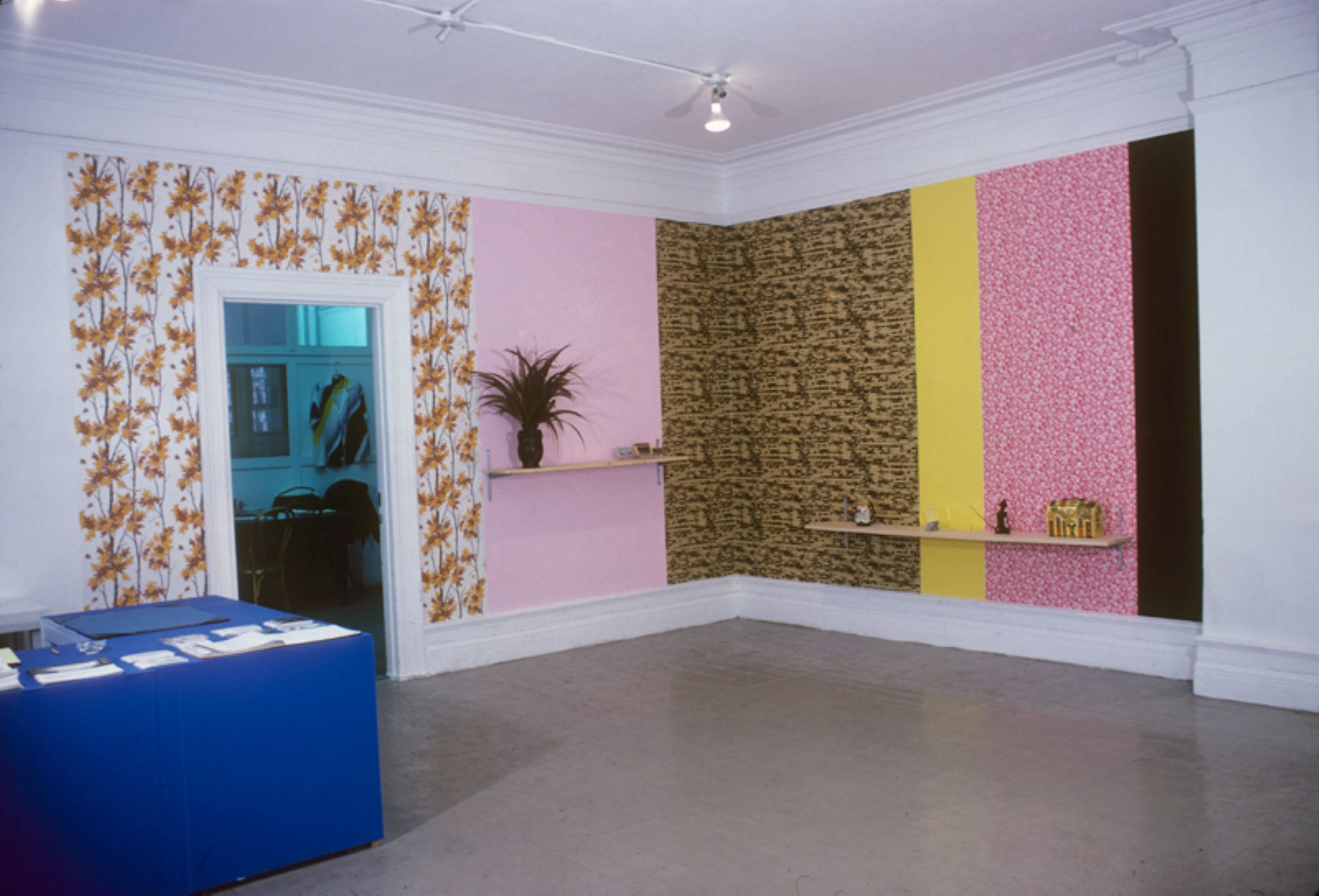 View of colorful panelled designed wall with shelves holding plants and other items. A blue desk with pamphlets on them is visible in the front left corner which is near a doorway into another room.