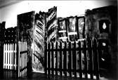 Black and white photograph of white picket fence with dark painted background behind it.