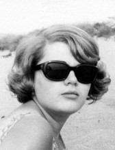 A black and white, portrait photograph of a woman at the beach. She wears sunglasses and has short, curled hair.